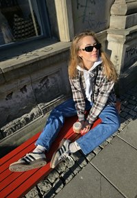 Sjekkert flanellskjorte lagd over en hvit hettegenser, blå jeans, grå joggesko med svarte striper, holder en kaffekopp, sittende på en rød benk.