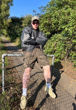 Chaqueta bomber negra, sudadera negra con capucha, pantalones cortos de camuflaje, zapatos de color claro. Sentado en una valla en un entorno natural al aire libre con vegetación.