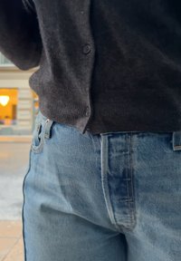 Personne portant un jean en denim bleu et un pull gris foncé boutonné, debout à l'extérieur près d'un bâtiment aux fenêtres éclairées.