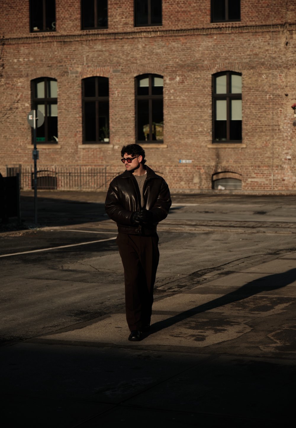 Homme portant une veste en cuir sombre, des gants et des lunettes de soleil, marchant dans la rue près d'un bâtiment en briques avec plusieurs fenêtres en arche, sous la lumière du soleil.