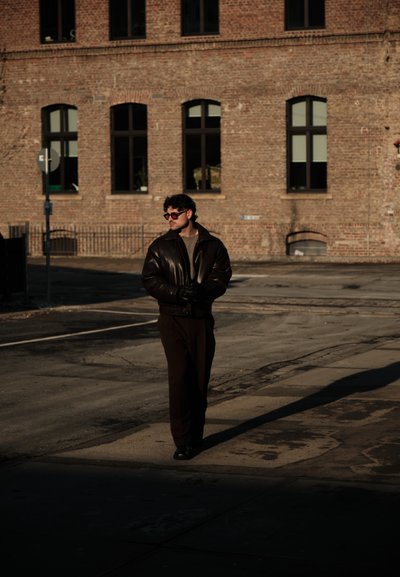 Hombre con chaqueta de cuero oscura, guantes y gafas de sol caminando por la calle cerca de un edificio de ladrillo con múltiples ventanas arqueadas bajo la luz del sol.
