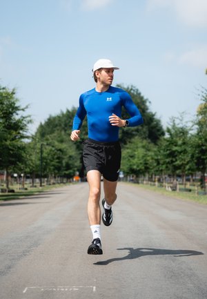 Atleta corriendo con una camiseta de manga larga azul y pantalones cortos negros, usando una gorra blanca y zapatillas de correr negras, en un camino bordeado de árboles.