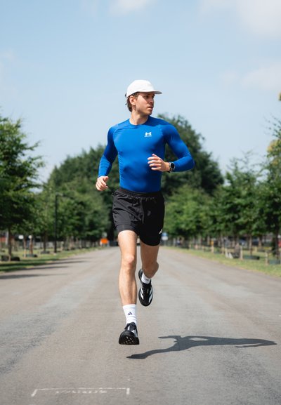 Atleta corriendo con una camiseta de manga larga azul y pantalones cortos negros, usando una gorra blanca y zapatillas de correr negras, en un camino bordeado de árboles.