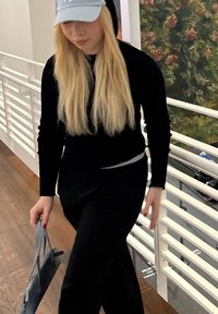 Jauna sieviete ar gariem blondiem matiem staigā iekštelpās, ģērbusies gaiši zilā cepurē un pilnīgi melnā apģērbā, turēdama nelielu pelēku rokassomu.