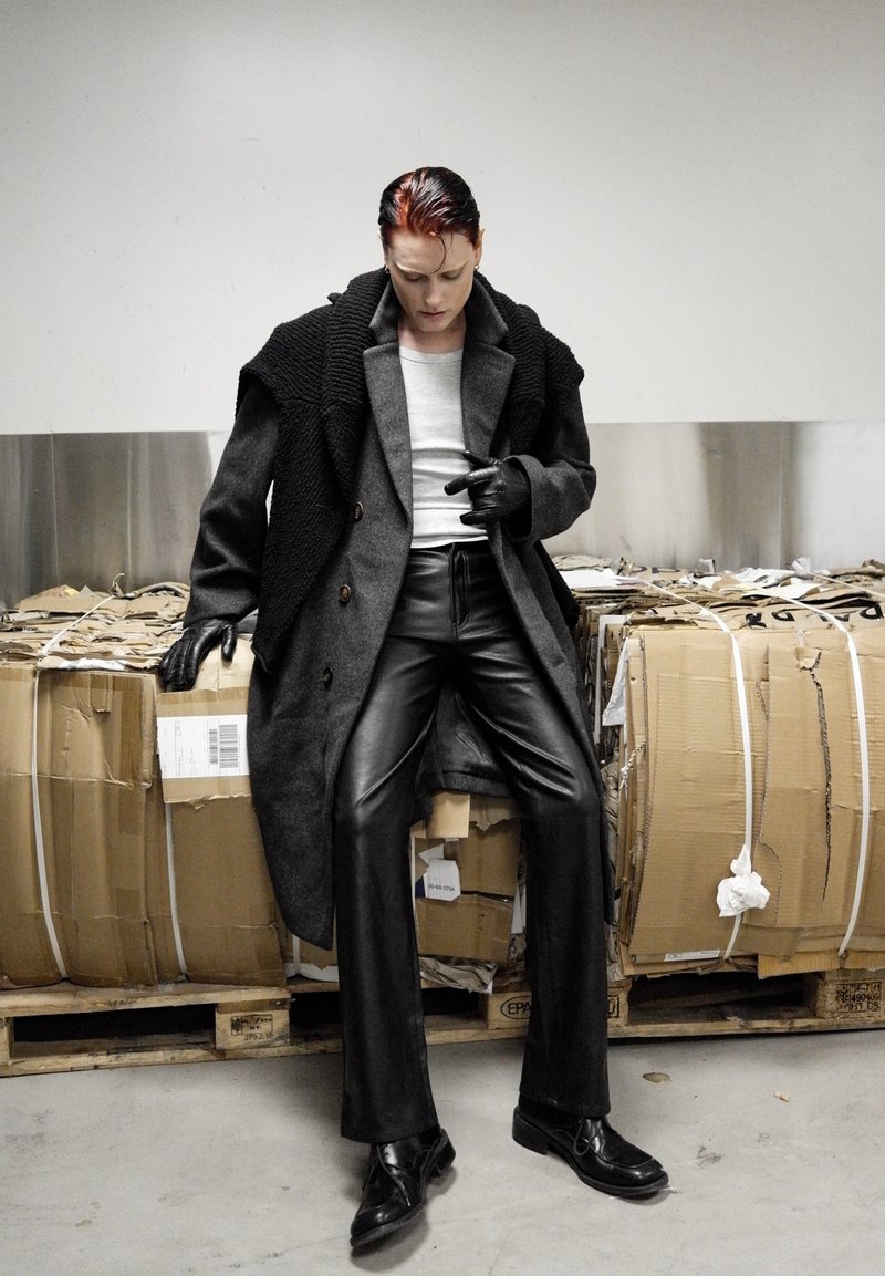 Pantalons en cuir noirs, manteau en laine grise avec des épaules texturées, débardeur gris, gants noirs et chaussures noires brillantes. Fond minimaliste avec du carton.
