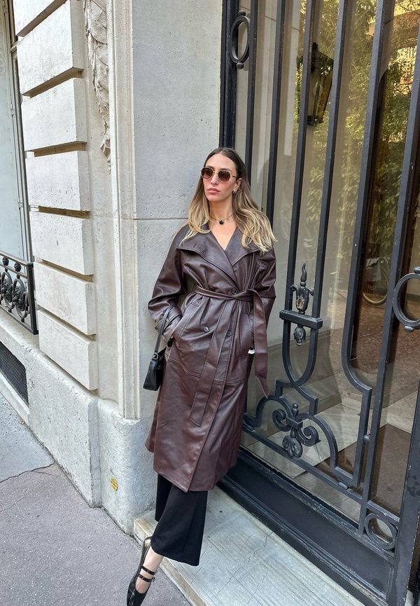 Mujer con abrigo trench de cuero marrón y gafas de sol apoyada en la esquina de un edificio de piedra cerca de una puerta negra de hierro forjado.