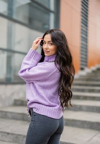 Pull tricoté violet avec un col haut, doté de poignets et d'un ourlet côtelés. Porté avec un jean gris foncé orné d'accents décoratifs sur le côté.