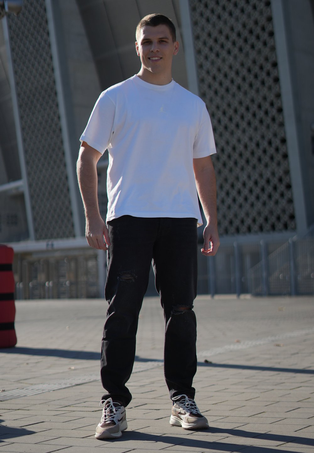 Bílá tričko s krátkým rukávem, černé džínové kalhoty s výřezem a světle barevné tenisky. Stojí venku s geometrickou strukturou v pozadí.