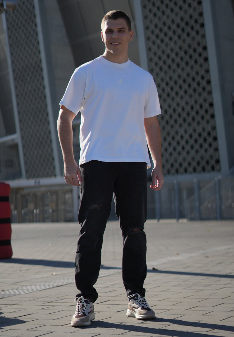 Hvid kortærmet t-shirt, sorte ripped jeans og lyse sneakers. Stående udendørs med en geometrisk struktur i baggrunden.