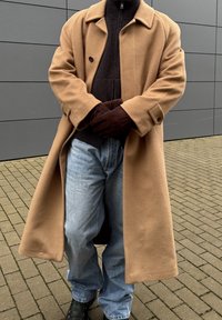 Μπεζ μάλλινο παλτό με μεγάλα πέτα και δύο κουμπιά, φορεμένο πάνω από σκούρο ριμπ πουλόβερ. Μπλε τζιν παντελόνι και καφέ γάντια ολοκληρώνουν την εμφάνιση.