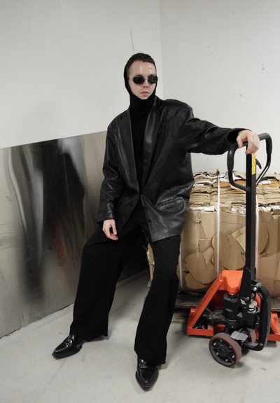Blazer de cuero negro oversize con un cuello alto, combinado con pantalones negros de pierna ancha. Accesorios incluyen gafas de sol oscuras. Entorno industrial.
