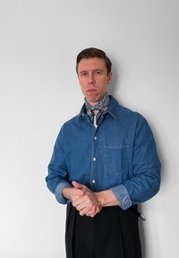 Άνδρας που φοράει μπλε πουκάμισο από τζιν και μαντήλι με πασλέ στο λαιμό, με τα χέρια ενωμένα, στέκεται μπροστά σε έναν απλό λευκό τοίχο.