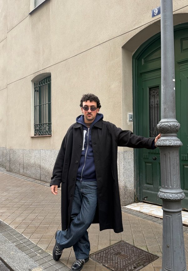 Uomo con occhiali da sole, cappotto nero e jeans appoggiato a un palo della luce su un marciapiede piastrellato vicino a una porta verde e una parete di edificio beige.