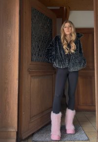 Crna jakna od umjetnog krzna, uske crne helanke i ružičaste čizme na petu s bijelim detaljima, postavljene uz drveni okvir vrata i pločice na podu.