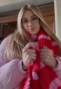 Vaaleanpunainen toppatakki, punavalkoraidallinen villahuivi, pitkät vaaleat hiukset, silmälasit, pitelee huivia molemmilla käsillä, taustalla puinen ovi.