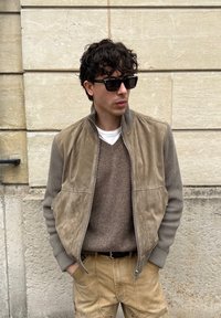 Νεαρός άνδρας που φοράει μαύρα γυαλιά ηλίου, καφέ μπουφάν με φερμουάρ, καφέ πουλόβερ και μπεζ παντελόνι, στέκεται μπροστά σε έναν τοίχο από πέτρα.