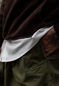 Sweat-shirt marron superposé sur une chemise blanche, associé à un short vert olive arborant un logo Nike en relief sur la poche.