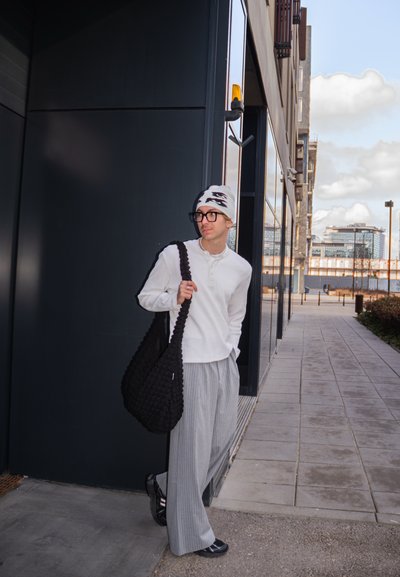 Joven con gorro, gafas, camiseta blanca de manga larga y pantalones anchos a rayas está cerca de la entrada de un edificio moderno llevando una gran bolsa negra.