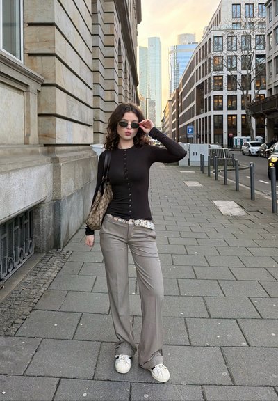Joven mujer con blusa negra y pantalones grises ajusta sus gafas de sol mientras está de pie en una acera urbana con edificios modernos y coches aparcados.