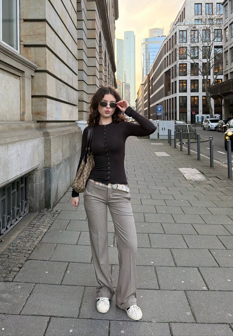 Jauna sieviete melnā topā un pelēkās biksēs noregulē saulesbrilles, stāvot pilsētas ietves malā pie modernām ēkām un novietotām automašīnām.