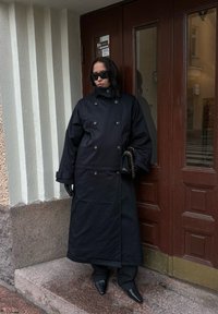 Black double-breasted long coat, oversized design, worn with black gloves and pointed-toe shoes, standing against a brown door.