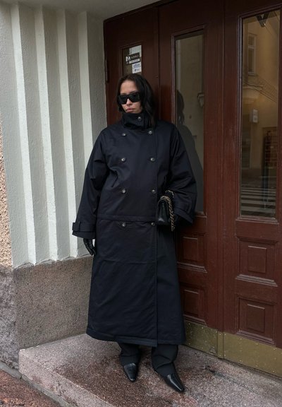 Abrigo largo negro con botones de doble hilera, combinado con guantes negros y zapatos de punta afilada. Está apoyado contra una puerta marrón con paneles de vidrio.