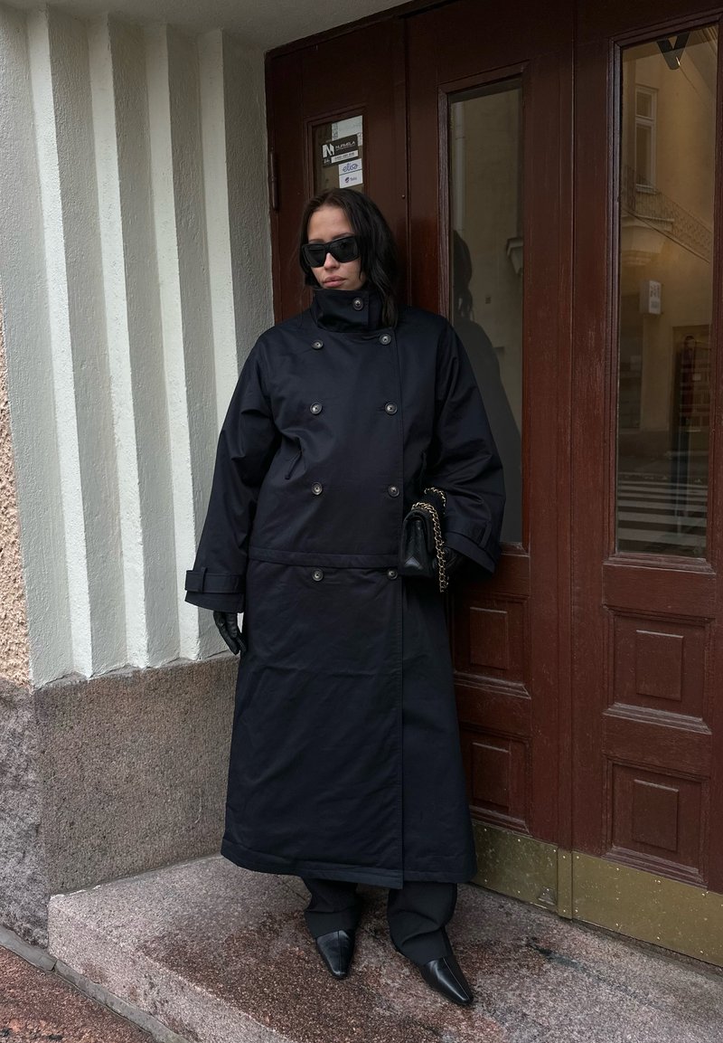 Czarny, dwurzędowy długi płaszcz o oversize'owym kroju, w komplecie z czarnymi rękawiczkami i butami z ostrym noskiem, stojący przed brązowymi drzwiami.