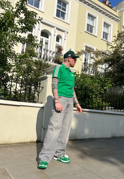 Polo de rayas verdes con blanco, jeans de pierna ancha grises y zapatillas verdes. El fondo presenta un edificio amarillo claro y vegetación.