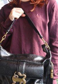 Personne en pull bordeaux tenant un sac à main en cuir noir Just Cavalli avec un emblème de serpent doré et un tire-zip.