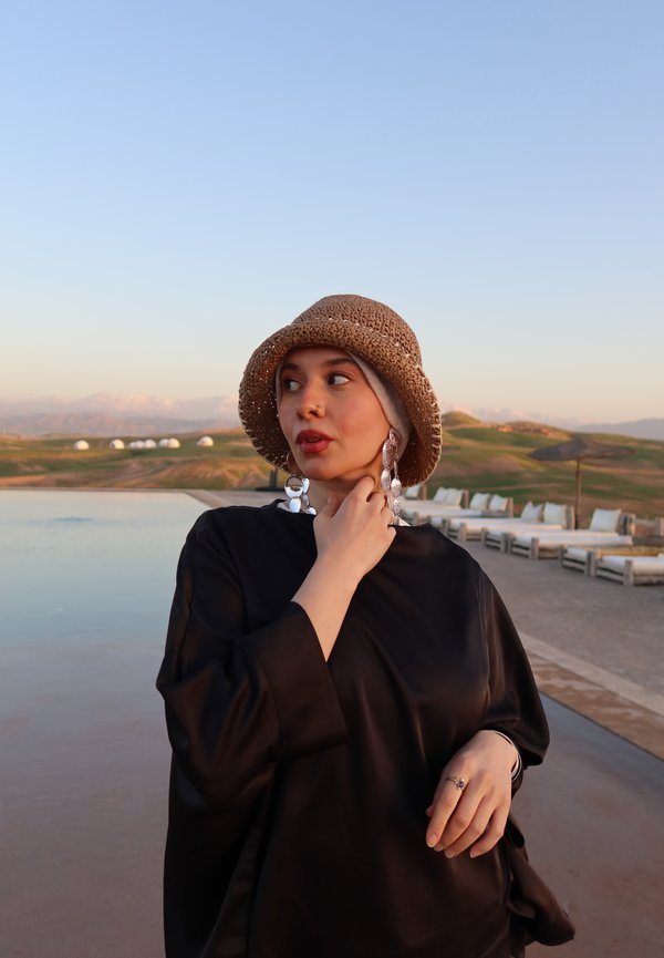 Femme portant un chapeau tressé marron, un haut noir et de grandes boucles d'oreilles en argent, debout près d'une piscine avec un paysage herbeux et un coin salon en arrière-plan.