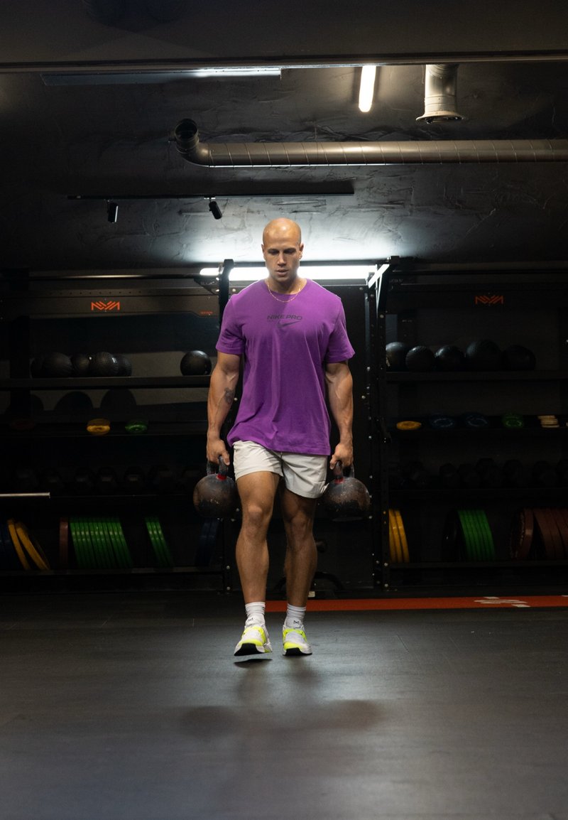 Figura maschile in una maglietta Nike Pro viola e pantaloni corti bianchi che porta due kettlebell, ambientato in una palestra con pesi sullo sfondo.