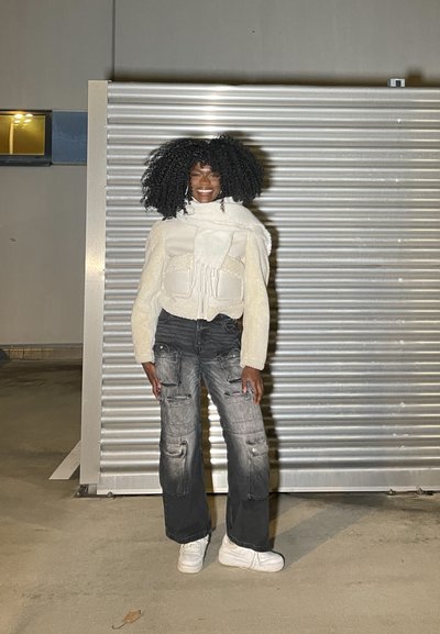 Chaqueta blanca texturizada corta con bolsillos de gran tamaño, combinada con pantalones cargo negros desgastados y holgados, y zapatillas blancas. El fondo presenta un panel de metal.