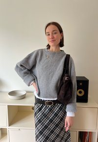 Jersey gris de punto acanalado, camisa blanca debajo, falda de cuadros en blanco y negro, cinturón negro con hebilla redonda y bolso de hombro marrón.