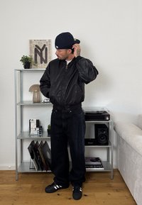 Jachetă neagră tip bomber cu fermoar, asociată cu blugi închiși la culoare și teniși negri Adidas. Ambient interior cu rafturi de cărți și decorațiuni.