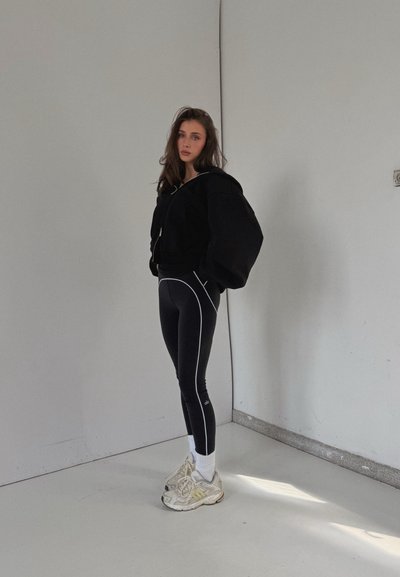 Sudadera negra corta, leggings negros de cintura alta con costuras blancas, calcetines blancos y zapatillas de color claro con suelas texturizadas. Fondo minimalista.