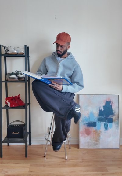 Hombre con gorra roja y sudadera gris con capucha sentado en un taburete, leyendo un libro, con un zapatero y una pintura abstracta cerca.