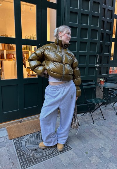 Chaqueta acolchada verde oliva con un acabado brillante, combinada con pantalones de chándal grises. El conjunto presenta un ajuste casual y botas color crema.