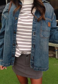 Femme portant une chemise à rayures, une veste en denim bleu et une jupe grise, debout sur une pelouse artificielle verte.