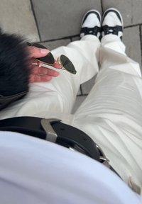 White wide-leg trousers paired with a black belt. A black furry sleeve is visible, with gold-framed sunglasses held in a hand. Black trainers are worn.