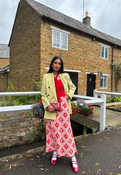 Blazer amarillo de doble botonadura, blusa roja con lazo, falda a rayas rojas y blancas, zapatos rojos, sosteniendo un pequeño bolso rojo. Fondo de casa de piedra.