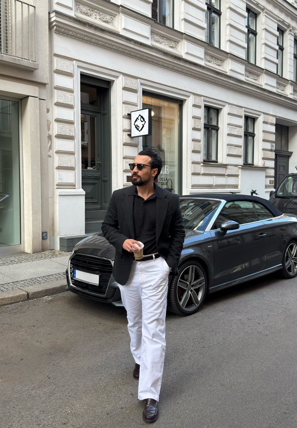 Hombre con gafas de sol, blazer negro y pantalones blancos sosteniendo una taza de café mientras camina junto a un coche descapotable negro en una calle de la ciudad.