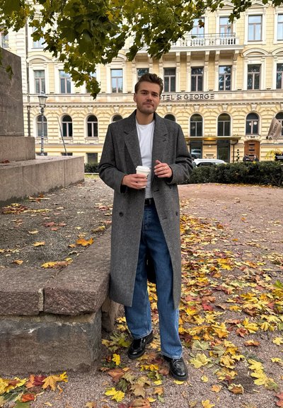 Abrigo de lana gris sobre una camiseta blanca, combinado con jeans azules y zapatos negros. Sujetando una taza de café, de pie en un camino cubierto de hojas de otoño.