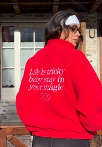 Femme portant une veste polaire rouge avec un texte blanc "La vie est compliquée bébé, reste dans ta magie", un bandeau blanc, des lunettes de soleil, debout en plein air près d'un bâtiment en pierre.