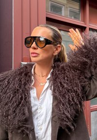 Femme aux cheveux blonds tirés en arrière, portant de grandes lunettes de soleil, un manteau foncé à poils longs et une chemise blanche, levant la main droite près d'une fenêtre de bâtiment.