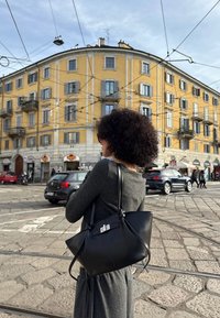Donna con capelli ricci e occhiali da sole standing su un angolo di strada in cobblestone, che porta una grande borsa nera, con un edificio giallo e auto sullo sfondo.