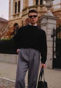 Černý pletený svetr, šedé plisované kalhoty, držící černou koženou tašku, stojící u ozdobné kamenné architektury s zelení v pozadí.