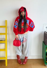Sweat à capuche rouge surdimensionné avec texte bleu et vert, pantalon de survêtement gris avec un motif rouge et bleu, sac à main rouge et bottes rouges brillantes.