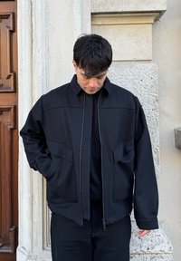 Jeune homme portant une veste noire et un pantalon, regardant vers le bas, debout contre un mur de pierre beige texturé près d'une porte en bois.