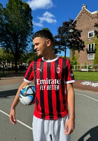 Camisa listrada em vermelho e preto com detalhes em branco, logótipo e texto "Emirates Fly Better"; calções cinzas; segurando uma bola de futebol preta e azul.