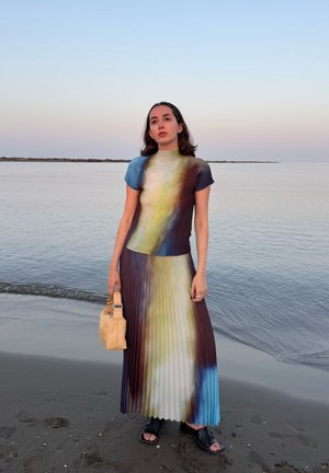 Vestido maxi plisado con un cuello alto, que presenta un degradado de tonos azules, verdes y marrones. Acompañado de un pequeño bolso beige y sandalias negras.